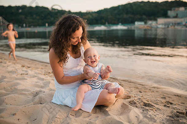 年轻的妈妈。坐着海滩一年婴儿儿子男孩拥抱微笑笑夏天一天快乐童年无忧无虑的游戏开放