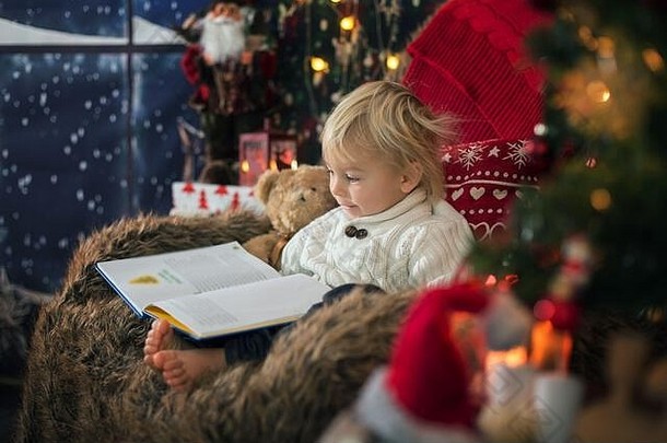 可爱的蹒跚学步的男孩坐着圣诞节树舒适的椅子阅读书圣诞节晚上