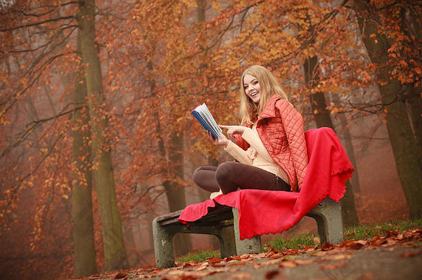 自然户外放松休闲文学概念夫人阅读蓝色的书年轻的金发女郎女孩秋天的森林坐着板凳上覆盖毯子