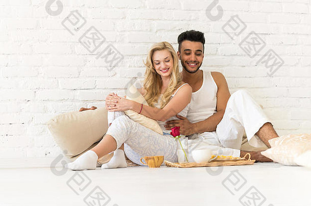 年轻的夫妇坐枕头地板上快乐拉美裔男人。女人早餐托盘情人卧室