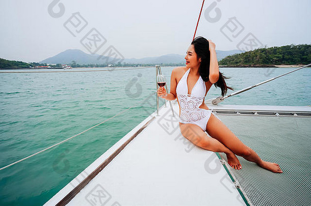 年轻的快乐亚洲女人美丽的脸享受持有葡萄酒杯坐着甲板航行游艇船放松比基尼