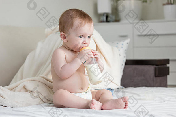 肖像可爱的婴儿男孩尿布坐着床上喝牛奶瓶