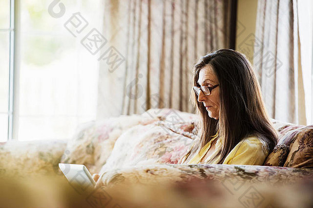高级女人长棕色（的）头发坐着沙发移动PC电脑穿阅读眼镜