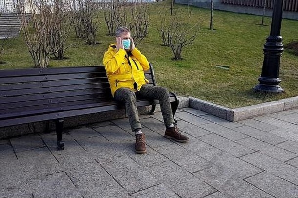 生病的男人。黄色的夹克穿医疗面具坐着公园板凳上