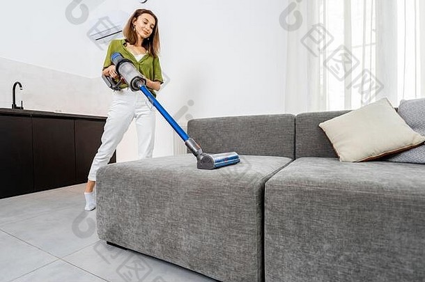 年轻的女人清洁沙发无线真空更清洁的现代白色生活房间概念容易清洁无线真空更清洁的
