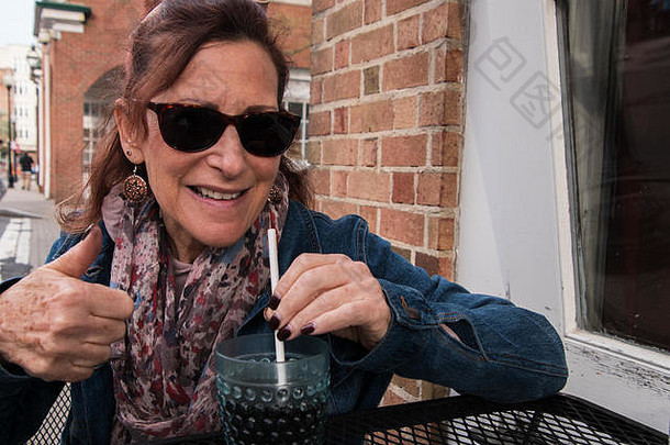 中间岁的婴儿潮女人穿太阳镜坐着户外餐厅表格街持有稻草花俏的塑料玻璃