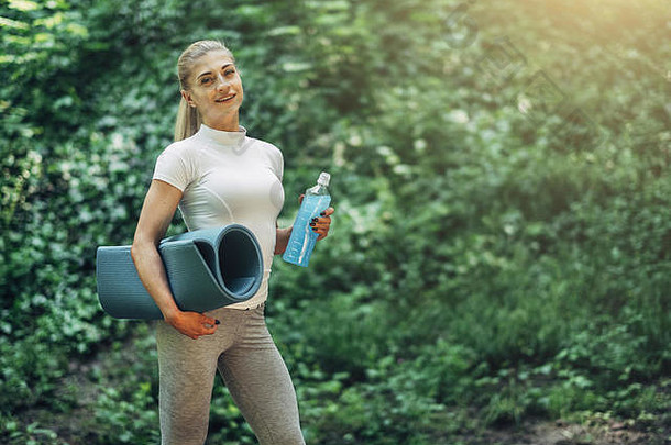 健身女人准备瑜伽早期早....公园穿时尚的体育运动装蓝色的瓶水瑜伽席健康的生活方式