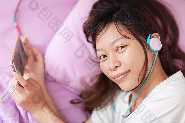 亚洲年轻的女孩看移动视频聪明的电话穿耳机说谎床上