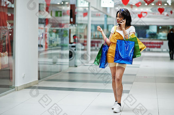 美丽的穿着考究的非洲式发型美国女人客户彩色的购物袋说话电话购物中心