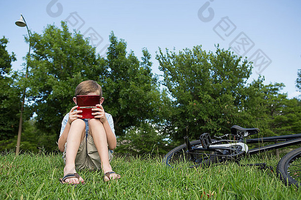 年轻的男孩坐着草玩手举行电子游戏