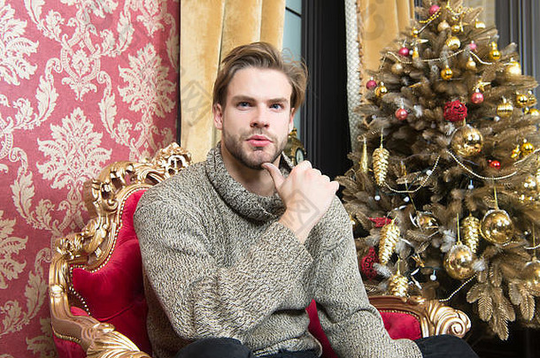冬天时尚风格概念男子气概毛衣圣诞节树生活房间假期准备庆祝活动男人。坐红色的扶手椅圣诞节树一年夏娃聚会，派对