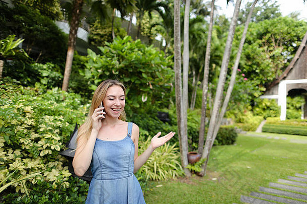年轻的高加索人女人会说话的智能手机花园树穿牛仔裤背心裙