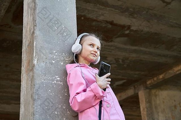 小高加索人女孩粉红色的斗篷听音乐耳机混凝土墙女孩持有智能手机手eenjoy音乐