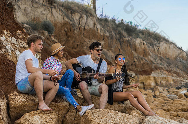 朋友坐着石头海滩男人。玩吉他