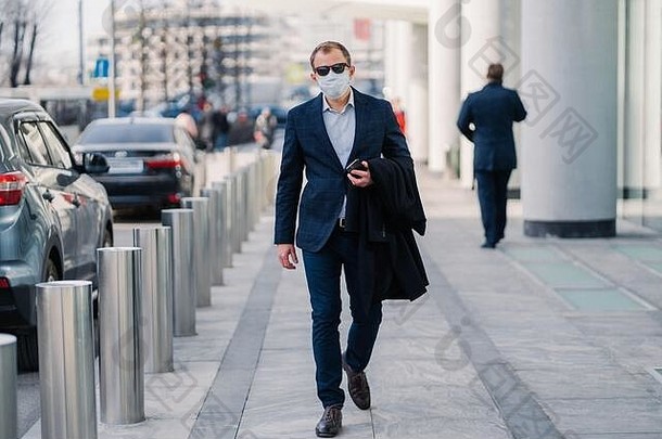 户外拍摄商人穿保护医疗面具走城市街穿着正式的衣服持有移动电话工作健康