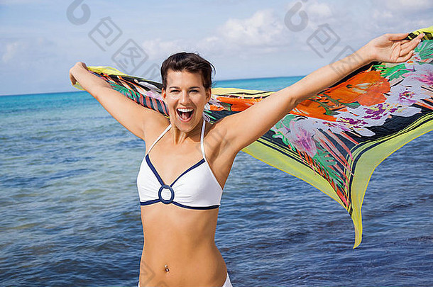 笑<strong>活泼</strong>的女人比基尼海边持有色彩鲜艳的有图案的围巾伸出的手颤振微风海洋背景