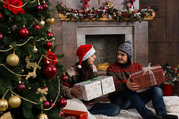 年轻的夫妇温暖的毛衣有趣的冬天帽子喜乐礼物坐着坐着地毯前面圣诞节树