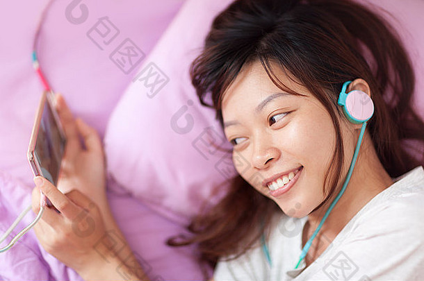 亚洲年轻的女孩看移动视频聪明的电话穿耳机说谎床上