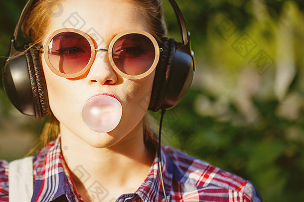 年轻的赶时髦的人女孩听音乐耳机夏天公园肖像特写镜头咀嚼口香糖温暖的爽肤水