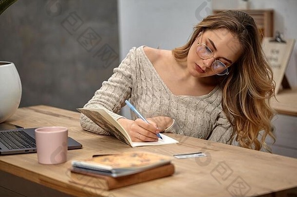 夫人博主眼镜米色毛衣坐着厨房表格写作笔记本移动PC书塑料卡粉红色的杯附近的关闭