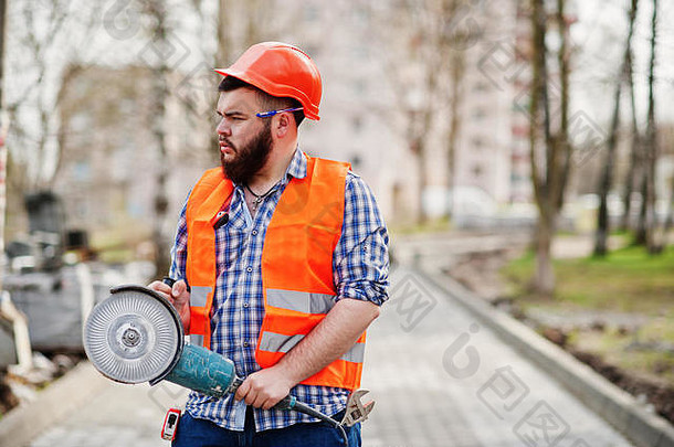 肖像残酷的胡子工人男人。西装建设工人安全橙色头盔人行道上角磨机手