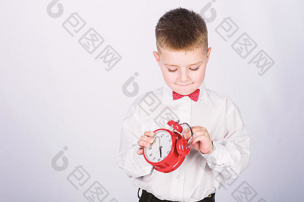 时间早....例程小学生报警时钟孩子可爱的男孩白色衬衫红色的弓领带开发纪律时间表时机集报警时钟孩子男孩持有红色的时钟