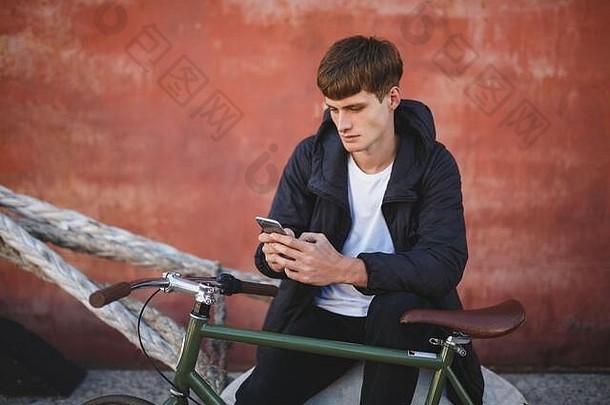 很<strong>酷</strong>的<strong>男孩</strong>棕色（的）头发坐着手机年轻的男人。夹克白色t恤坐着移动电话手自行车