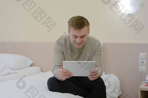 年轻的男人。未来酒店房间坐着床上视频闲谈，聊天平板电脑