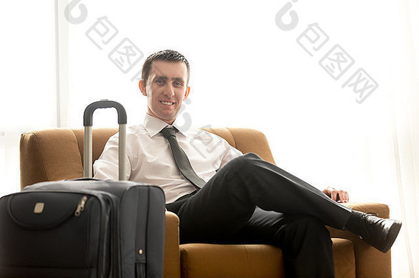 肖像英俊的微笑年轻的业务男人。行李坐着扶手椅酒店房间