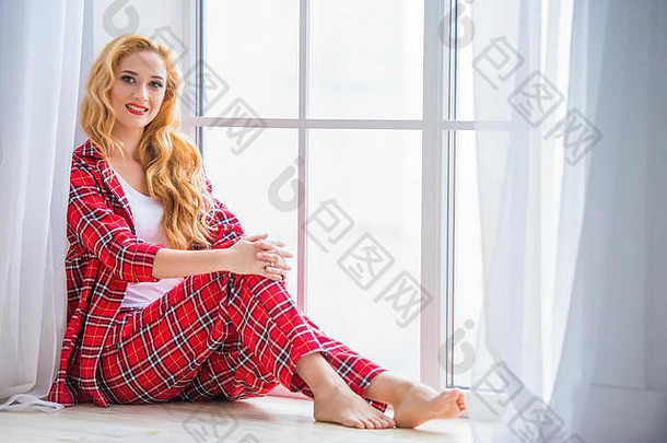 美丽的金发女郎女人红色的网纹睡衣坐在窗口演示了舒适的首页衣服
