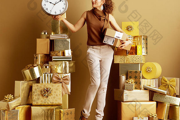 完整的长度肖像微笑年轻的女人黄金米色裤子棕色（的）上衣包裹时钟桩金礼物前面