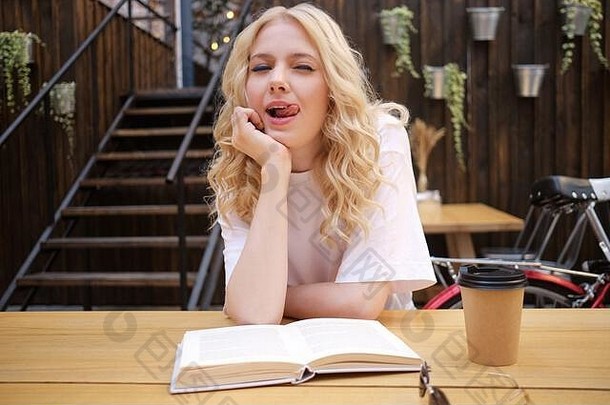 有吸引力的金发碧眼的女孩开玩笑地显示舌头相机坐着书咖啡院子里城市咖啡馆
