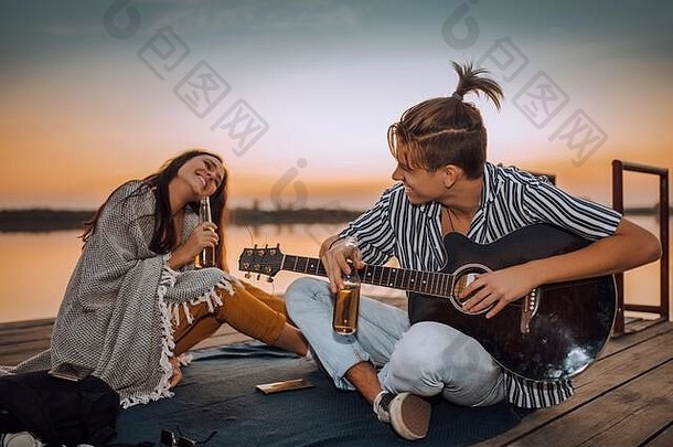快乐快乐的年轻的夫妇喝<strong>啤酒</strong>玩吉他唱歌坐着木浮筒河