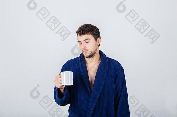 肖像伤心困了年轻的男人。混乱的头发穿蓝色的浴袍喝咖啡杯孤立的白色背景